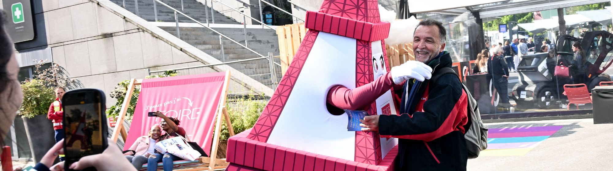 Un visiteur prend une photo avec la mascotte Tour Eiffel à la Foire de Paris pendant qu’un autre groupe se prend en selfie sur un transat