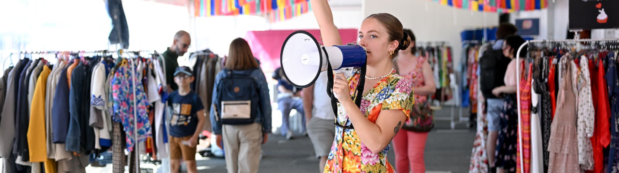 Animation festive au cœur du marché de la seconde main à foire de paris, avec une animatrice au mégaphone dans une allée colorée de vêtements vintage.