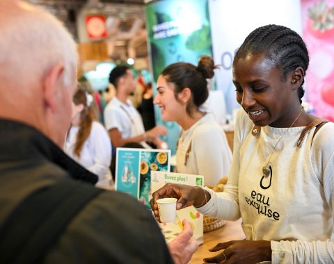 Animatrice servant un échantillon de boisson à un visiteur au stand 'eau exquise' lors de la Foire de Paris